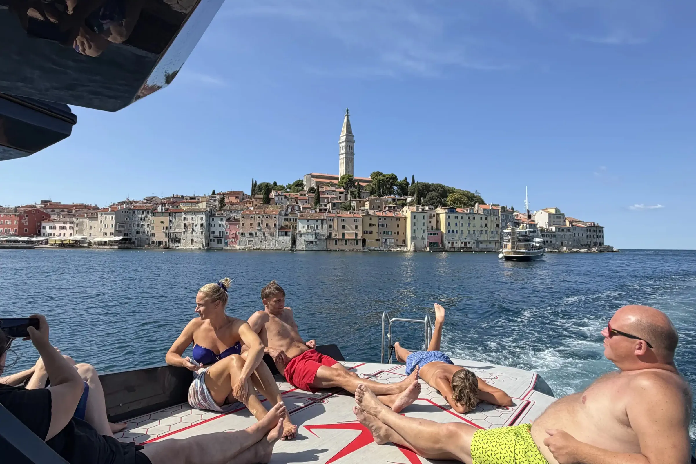 Gosti leže na pramčanoj palubi dok brod stoji pred starim gradom Rovinjem sa zvonikom na vrhu brijega
